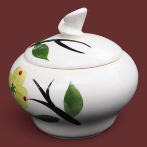 May include: A white ceramic sugar bowl with a lid featuring a sculptural handle. The bowl is decorated with hand-painted black branches, green leaves, and a yellow flower with red centers. The smooth, glossy finish covers the entire rounded vessel.