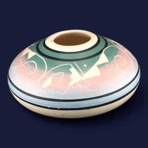 May include: A low-profile, rounded ceramic vessel featuring a wide opening at the top. The exterior displays a gradient of muted green, soft pink, and light blue, accented by thin black horizontal lines and white etched geometric patterns.