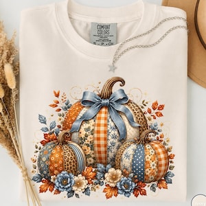 May include: An ivory t-shirt featuring a graphic of three pumpkins with varied patterns including leopard print, gingham, and floral designs. The central pumpkin is adorned with a large blue ribbon. The design includes autumn leaves and flowers.