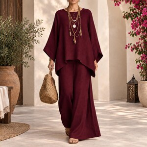 May include: A deep burgundy two-piece set consisting of a loose-fitting long-sleeve top with wide sleeves and matching wide-leg trousers. The outfit is paired with a textured tan woven handbag and a layered gold-toned pendant necklace.