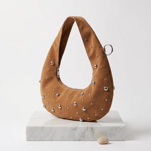 May include: A crescent-shaped tan suede shoulder bag featuring a smooth texture and scattered silver-toned metal studs of varying sizes. The bag includes a single integrated handle, a silver zipper closure, and a circular silver metal ring attachment.