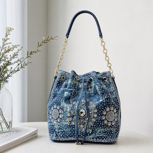 May include: A blue acid-wash denim bucket bag featuring a drawstring closure, gold-tone chain shoulder strap, and intricate circular patterns made of clear rhinestones. The bag is embellished with scattered crystals across the entire surface.