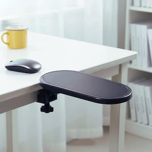May include: A black ergonomic armrest support attachment for a desk, featuring an oval-shaped padded surface and a sturdy screw-clamp mechanism for secure mounting to the edge of a table, positioned near a computer mouse and a yellow mug.