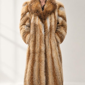 Può includere: Un cappotto lungo a maniche lunghe caratterizzato da un esterno in pelliccia sintetica spessa e testurizzata con strisce verticali nei toni del marrone, beige e bianco. Il capo presenta un ampio colletto a revers ed è indossato sopra pantaloni e stivali neri.