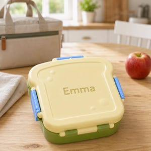 Peut inclure: Une boîte à déjeuner carrée jaune clair en forme de tranche de pain avec le prénom Emma gravé sur le couvercle. Le récipient présente des clips de verrouillage bleus sur les côtés et une base vert foncé pour conserver les aliments.