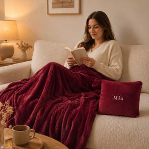 May include: A deep burgundy plush throw blanket with a matching square velvet pillow featuring the embroidered name Mia in white thread. The soft, textured fabric of the blanket drapes over the legs, while the pillow rests on a cream sofa.