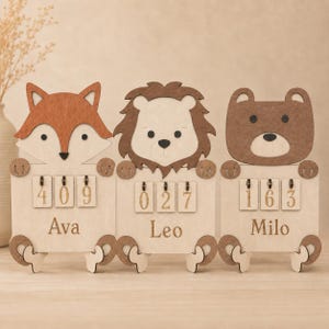 Peut inclure: Trois plaques en bois en forme d'animaux représentant un renard, un lion et un ours. Chaque plaque comporte trois tuiles numérotées à bascule affichant 409, 027 et 163, avec les prénoms Ava, Leo et Milo gravés sous les chiffres.