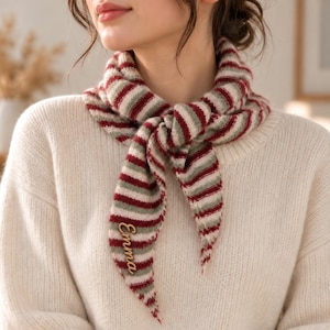 May include: A knitted triangular scarf featuring horizontal stripes in shades of dark red, sage green, and cream. The scarf is tied at the neck and includes the name Emma embroidered in a cursive script on one of the pointed ends.