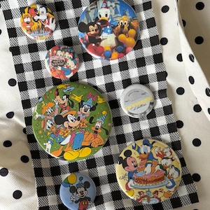 May include: A collection of seven circular pin-back buttons featuring various Disney characters including Mickey Mouse, Donald Duck, Goofy, and Pluto. The buttons display colorful illustrations of birthday celebrations and anniversaries, with one silver button labeled Small Shop Buttons.