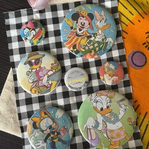 May include: A collection of six circular pin-back buttons featuring various Disney characters including Minnie Mouse and Daisy Duck in different outfits. A small silver metal tin with the text Small Shop Taymode Buttons is in the center.