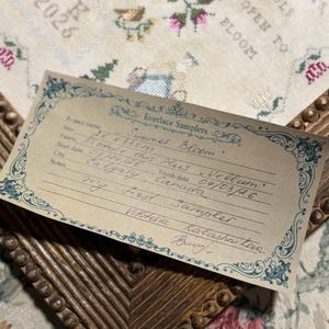 Puede incluir: Una tarjeta de proyecto rectangular Everlace Samplers con detalles escritos a mano que incluyen Summer Bloom, 26 x 33cm, y Calgary, Canada, sobre un marco de madera encima de un bordado de punto de cruz con motivos florales y el texto HEARTS SOULS OPEN TO BLOOM.