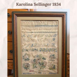 Könnte beinhalten: Ein gerahmter Kreuzstich-Sampler mit dem Titel Everlace Samplers Karolina Sellinger 1834. Das Stück zeigt Reihen von Buchstaben, Blumenmotiven und dekorativen Rändern, die mit blauem und grünem Garn auf hellem Stoff gestickt wurden.