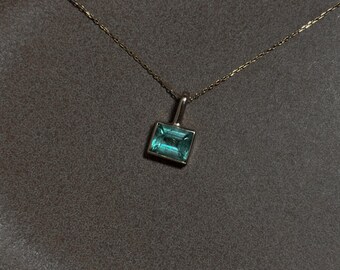 May include: A delicate gold chain necklace featuring a single rectangular, emerald-cut teal gemstone pendant set in a simple gold frame. The pendant hangs centrally from the thin, polished gold metal chain, showcasing a clear, vibrant blue-green stone.