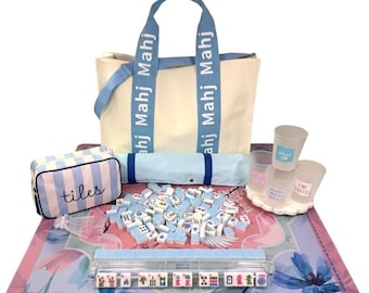 May include: A mahjong set featuring a cream-colored tote bag with light blue straps labeled Mahj, a striped pouch labeled tiles, a pink floral play mat, a clear tile rack, a pile of light blue and white tiles, and three small cups labeled Mahj.