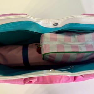 May include: A tote bag with a white exterior and bright pink base, featuring a vibrant turquoise interior lining. Inside the bag sits a rectangular zippered pouch with a light pink and white vertical striped pattern and a green trim border.