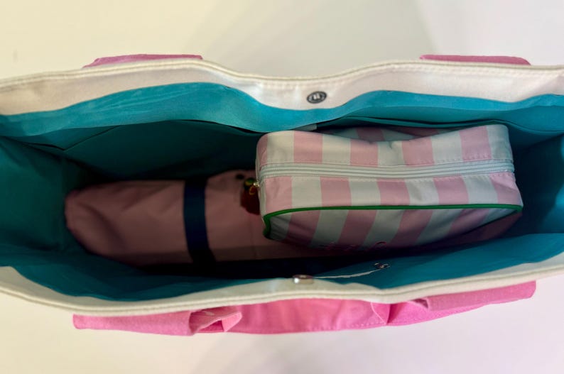May include: A tote bag with a white exterior and bright pink base, featuring a vibrant turquoise interior lining. Inside the bag sits a rectangular zippered pouch with a light pink and white vertical striped pattern and a green trim border.