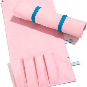 May include: A light pink rectangular organizer mat featuring four vertical pockets along the bottom edge. The mat includes two bright blue elastic bands for securing it in a rolled position, and metal grommets at the top corners for hanging.