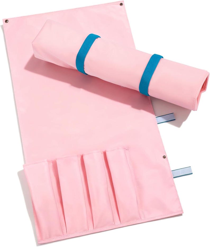 May include: A light pink rectangular organizer mat featuring four vertical pockets along the bottom edge. The mat includes two bright blue elastic bands for securing it in a rolled position, and metal grommets at the top corners for hanging.