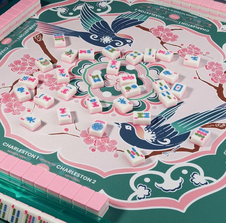 May include: A mahjong set featuring a green and pink play mat decorated with illustrations of birds and cherry blossoms. The mat includes text for Charleston rules. Scattered white tiles with colorful symbols and a Joker tile are on the mat.