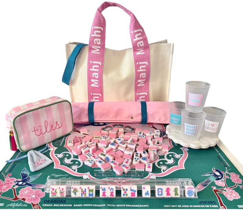 May include: A Mah Jong set featuring a cream canvas tote bag with pink straps labeled Mahj, a pink striped tile pouch, a green play mat with floral patterns, a clear tile rack, pink and white game tiles, and four frosted plastic cups with labels.