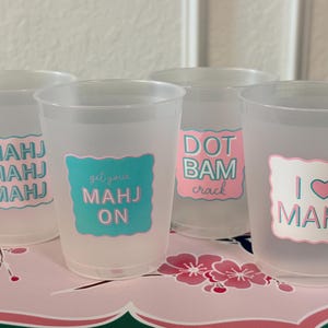 May include: Four frosted plastic cups featuring colorful decals with text. The first cup says MAHJ MAHJ MAHJ, the second says get your MAHJ ON, the third says DOT BAM crack, and the fourth says I heart MAHJ. The designs use pink and teal scalloped borders.