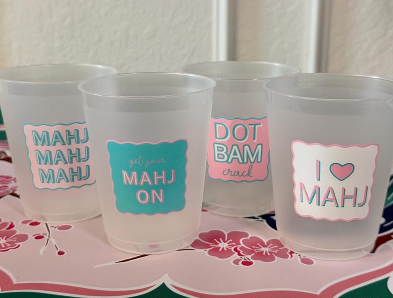 May include: Four frosted plastic cups featuring colorful decals with text. The first cup says MAHJ MAHJ MAHJ, the second says get your MAHJ ON, the third says DOT BAM crack, and the fourth says I heart MAHJ. The designs use pink and teal scalloped borders.