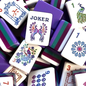 May include: A collection of Mahjong tiles featuring white faces with colorful illustrations of peacocks, floral patterns, and the word JOKER. The tiles have thick, layered sides in alternating bands of purple, dark green, and white plastic material.