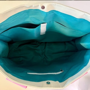 May include: A large tote bag with a cream-colored exterior and a vibrant turquoise interior lining. The bag features a magnetic snap closure at the top, an open interior pocket, and a zippered pocket on the inside wall for secure storage.