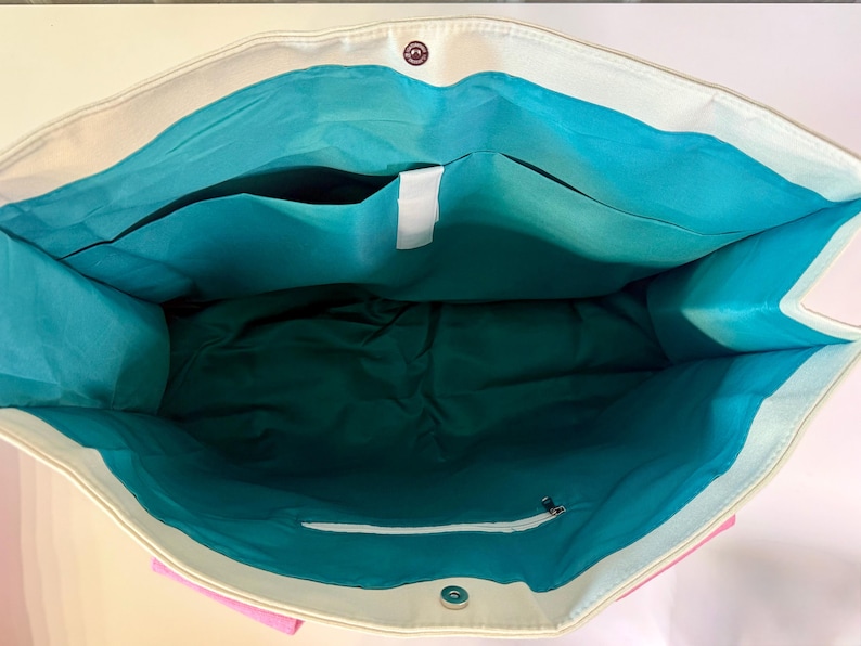 May include: A large tote bag with a cream-colored exterior and a vibrant turquoise interior lining. The bag features a magnetic snap closure at the top, an open interior pocket, and a zippered pocket on the inside wall for secure storage.