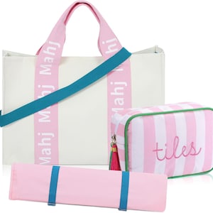 May include: A white canvas tote bag with pink straps featuring the text Mahj Mahj, a teal shoulder strap, a pink and white striped rectangular pouch with the word tiles embroidered in pink, and a long pink roll with two teal elastic bands.