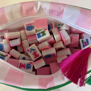May include: A zippered pouch with a pink and white striped pattern containing a collection of small, rectangular mahjong tiles. The tiles feature various pink, blue, and green symbols and characters. A bright pink and a green tassel hang from the zipper.