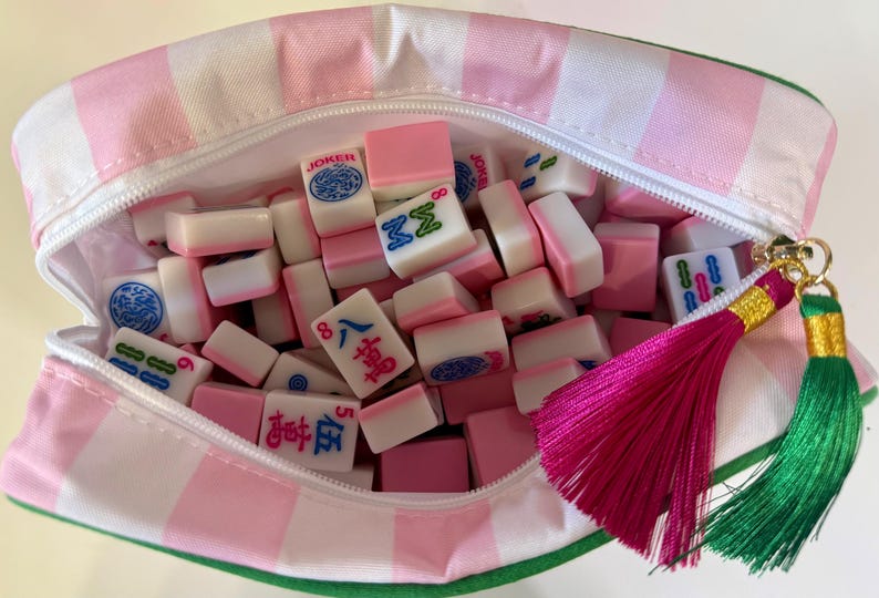 May include: A zippered pouch with a pink and white striped pattern containing a collection of small, rectangular mahjong tiles. The tiles feature various pink, blue, and green symbols and characters. A bright pink and a green tassel hang from the zipper.