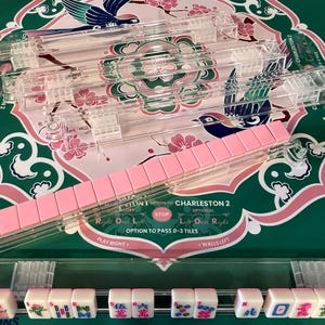 May include: A set of clear acrylic Mahjong tile racks, with one rack holding a row of pink-backed tiles and another rack displaying white tiles with colorful character, bamboo, and dragon designs, featuring text including Charleston 2.