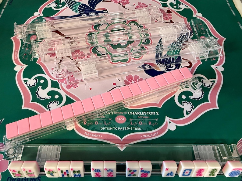 May include: A set of clear acrylic Mahjong tile racks, with one rack holding a row of pink-backed tiles and another rack displaying white tiles with colorful character, bamboo, and dragon designs, featuring text including Charleston 2.