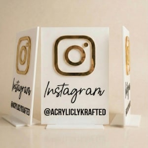 Może przedstawiać: Biały prostokątny szyld akrylowy ze złotym logo Instagrama u góry, słowem Instagram napisanym czarną kursywą oraz tekstem @ACRYLICLYKRAFTED czarnymi drukowanymi literami u dołu, umieszczony na przezroczystej akrylowej podstawie.