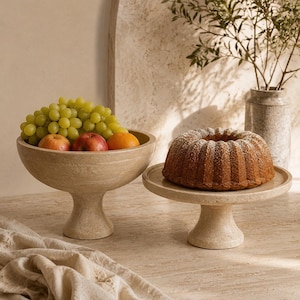 May include: A beige travertine pedestal bowl filled with green grapes, red apples, and an orange, alongside a matching flat travertine cake stand holding a golden-brown bundt cake dusted with white powdered sugar, set on a stone surface.