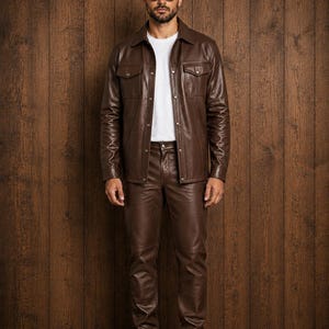 Könnte beinhalten: Eine dunkelbraune Lederhemdjacke mit zwei Brusttaschen und Metalldruckknöpfen, getragen über einem schlichten weißen T-Shirt. Das Outfit umfasst eine passende dunkelbraune Lederhose, schwarze Lederstiefel und eine dunkel getönte Sonnenbrille.