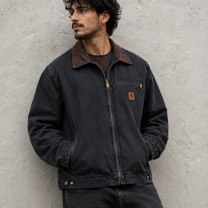 Könnte beinhalten: Eine anthrazitfarbene Arbeitsjacke aus Canvas mit dunkelbraunem Cordkragen, durchgehendem Metallreißverschluss und einem kleinen rechteckigen Carhartt-Markenaufnäher auf der linken Brusttasche sowie zwei seitlichen Eingrifftaschen und einer Brusttasche.