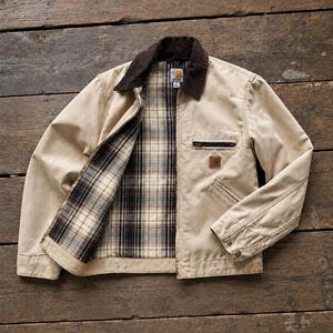 Könnte beinhalten: Eine beigefarbene Canvas-Jacke mit dunkelbraunem Cordkragen, Reißverschluss vorne und einem karierten Flanellfutter in Braun-, Schwarz- und Cremetönen. Die Jacke verfügt über eine Brusttasche mit Reißverschluss und ein quadratisches Carhartt-Logo.