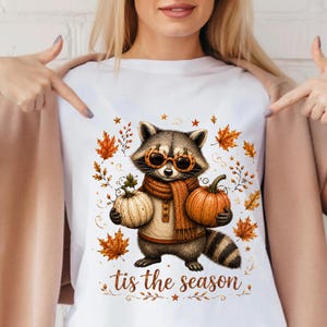 May include: A white t-shirt featuring a graphic of a raccoon wearing a tan sweater, a brown scarf, and pumpkin-shaped glasses. The raccoon holds a white pumpkin and an orange pumpkin, surrounded by autumn leaves and the text "tis the season".