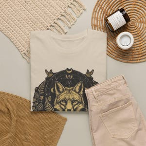 Può includere: Una t-shirt beige con una stampa grafica scura raffigurante un muso di volpe al centro di un design celeste circolare. L'illustrazione include dettagli intricati di felci, pigne, uccelli e fasi lunari disposti in uno schema simmetrico.