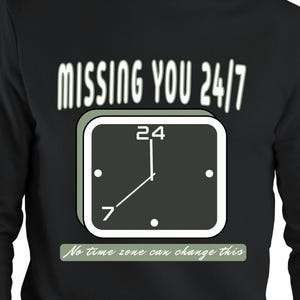 Może przedstawiać: Czarna bluza z długim rękawem z grafiką kwadratowego zegara, napisem MISSING YOU 24/7 powyżej oraz napisem No time zone can change this na jasnozielonym prostokątnym tle pod ilustracją tarczy zegara.