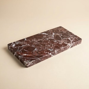 May include: A rectangular serving board crafted from deep reddish-brown marble, featuring a polished surface with prominent white and lighter brown veining throughout the stone. The edges are straight and defined, showcasing the natural pattern.
