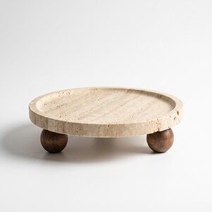 May include: A circular beige travertine stone tray with a porous, textured surface and a raised rim. The tray is supported by three dark brown spherical wooden feet, providing a stable base for the flat, round stone platform.
