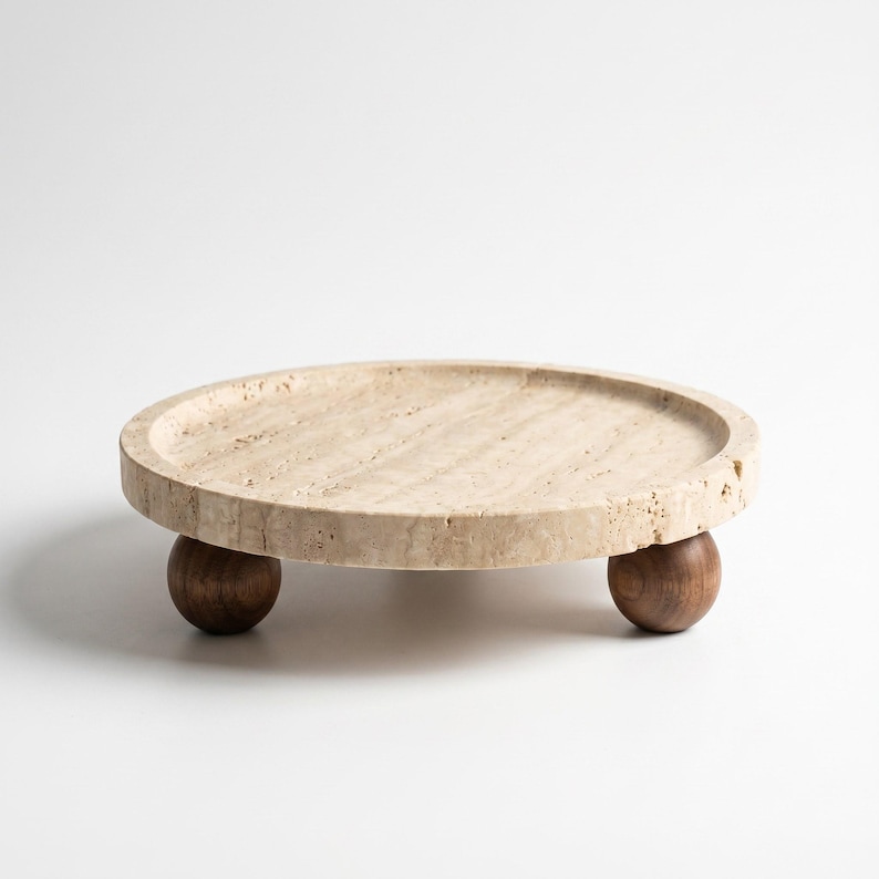 May include: A circular beige travertine stone tray with a porous, textured surface and a raised rim. The tray is supported by three dark brown spherical wooden feet, providing a stable base for the flat, round stone platform.