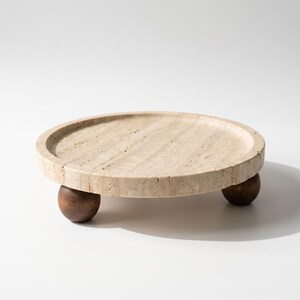 May include: A circular beige travertine stone tray with a natural porous texture and a raised rim. The tray is elevated by three dark brown spherical wooden feet, providing a stable base for this decorative stone display piece.