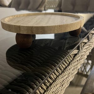 May include: A circular beige travertine stone tray with a porous, textured surface and a raised rim. The tray is elevated by three small, spherical dark brown wooden feet positioned evenly underneath the base to provide stability and height.