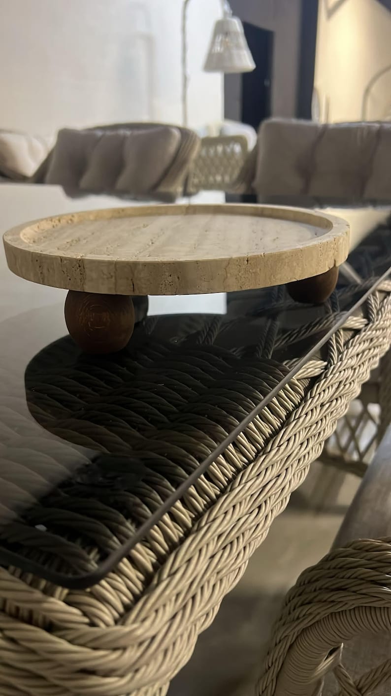 May include: A circular beige travertine stone tray with a porous, textured surface and a raised rim. The tray is elevated by three small, spherical dark brown wooden feet positioned evenly underneath the base to provide stability and height.