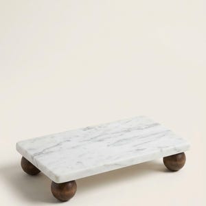 Puede incluir: Una losa rectangular de mármol blanco con vetas grises, elevada por cuatro pies esféricos de madera de color marrón oscuro. La superficie es lisa y pulida, con un patrón de piedra natural que contrasta con los soportes redondeados de madera.