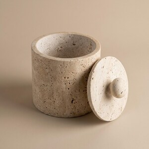 Peut inclure: Un pot cylindrique en pierre de travertin beige avec une surface poreuse et texturée, accompagné d'un couvercle circulaire assorti. Le couvercle est doté d'une petite poignée sphérique sur le dessus. L'intérieur du pot est creux et naturel.